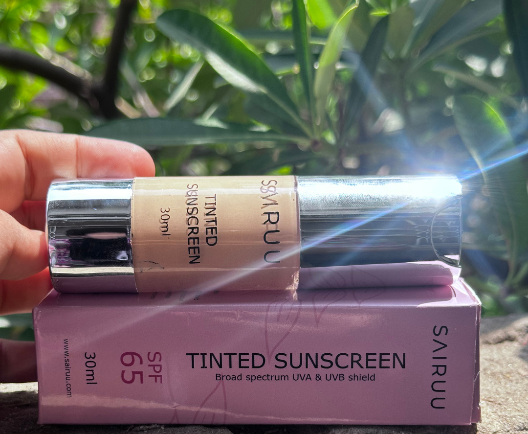 A hand holds a tube of SAIRUUN tinted sunscreen over its pink box, set outdoors on a stone surface with green foliage in the background.