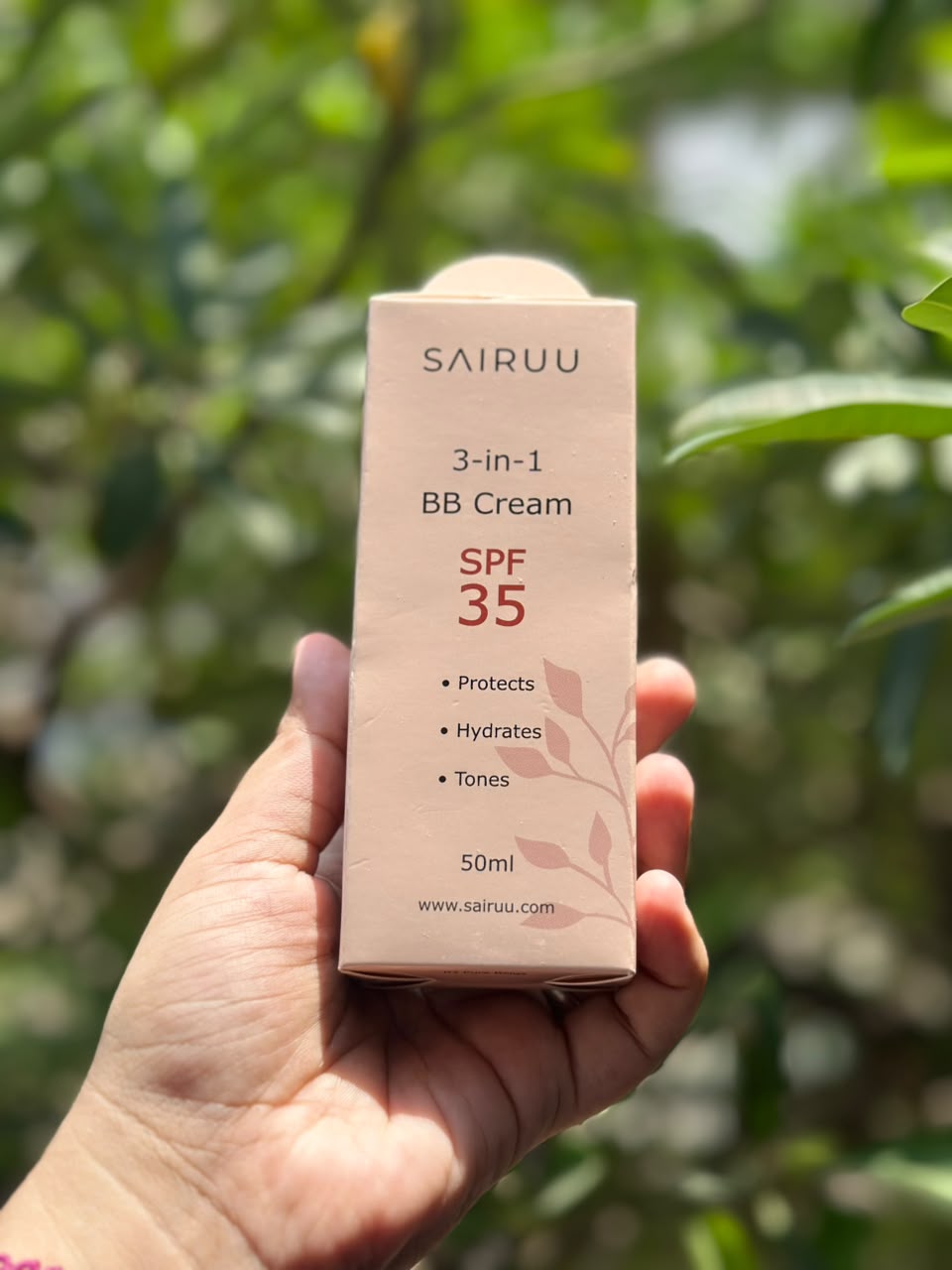A hand holding a pink box of Sairuu 3-in-1 BB Cream SPF 35, with a blurred green outdoor background.