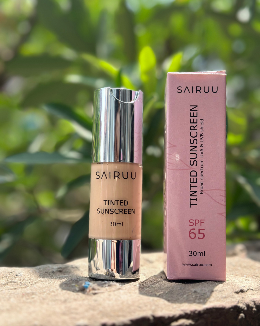 SAIRUU Tinted Sunscreen SPF 65 in a 30ml bottle with a reflective silver cap, standing next to its pink packaging box on a stone surface with a blurred green garden background.
